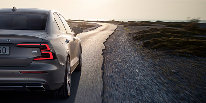 View showing rear of a grey Volvo saloon driving along a semi-metalled road